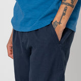 Person wearing a blue shirt and dark pants with tattoos on arms against a plain background