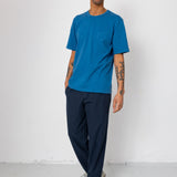 Man wearing a blue t-shirt and navy pants standing against a white wall.