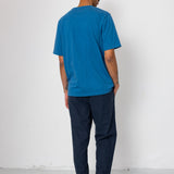 Person wearing a blue t-shirt and dark pants standing against a white wall.