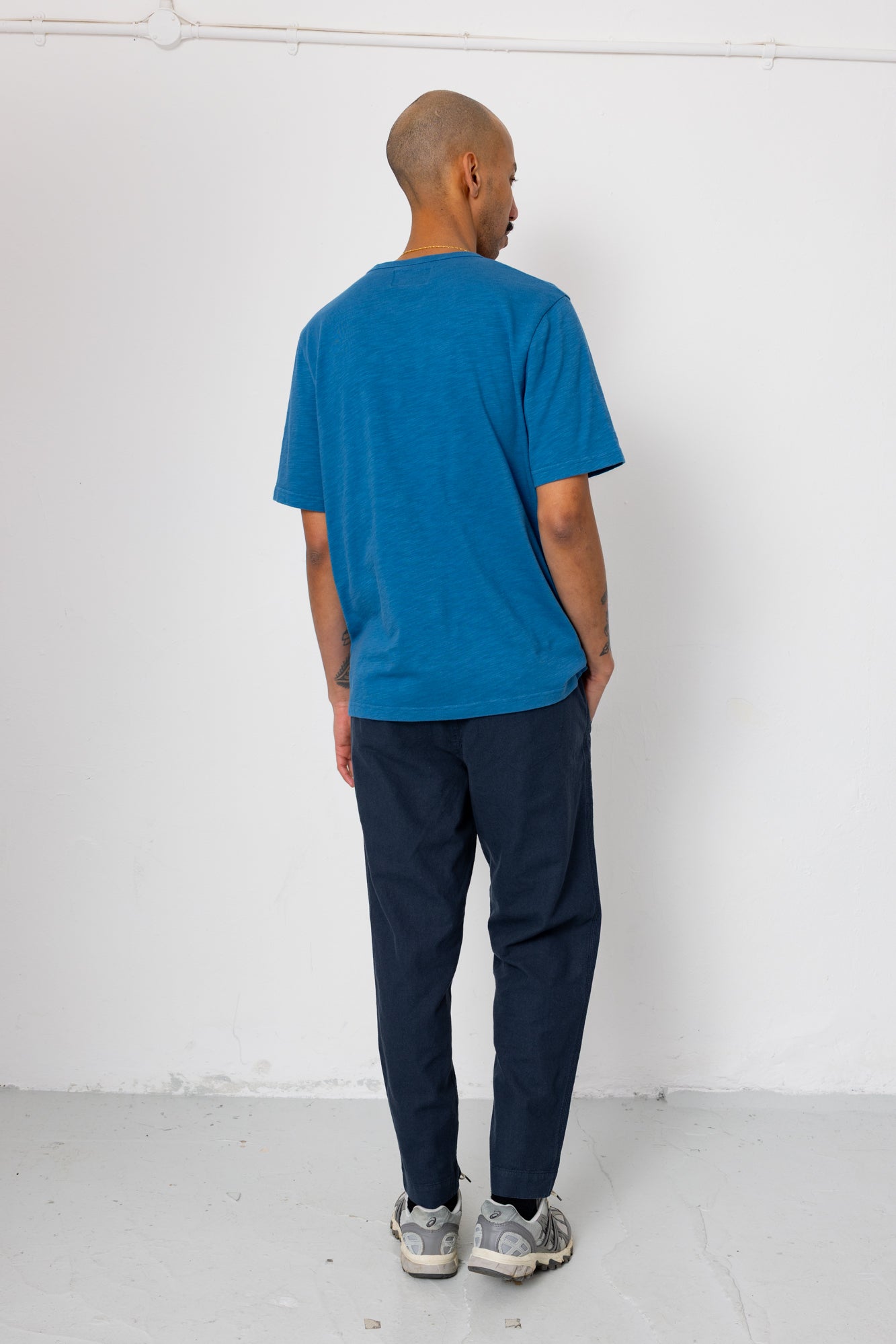 Person wearing a blue t-shirt and dark pants standing against a white wall.