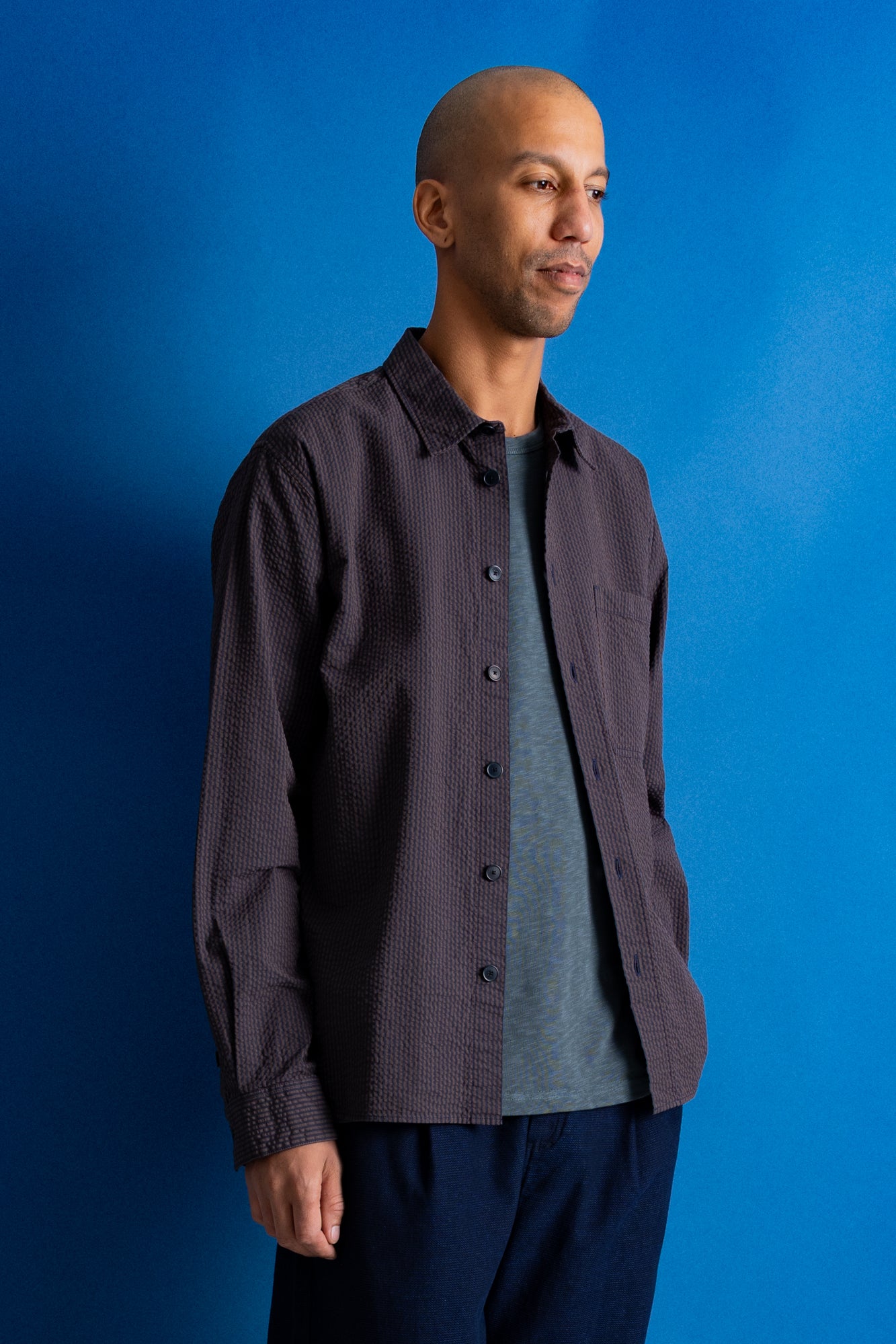 A man wearing a textured dark gray button-down shirt over a heather gray t-shirt and navy pants, standing against a solid blue studio background.