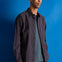 A man wearing a textured dark gray button-down shirt over a heather gray t-shirt and navy pants, standing against a solid blue studio background.