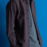 A man wearing a dark textured button-up shirt over a heather gray t-shirt and navy pants, posed against a solid blue studio background.