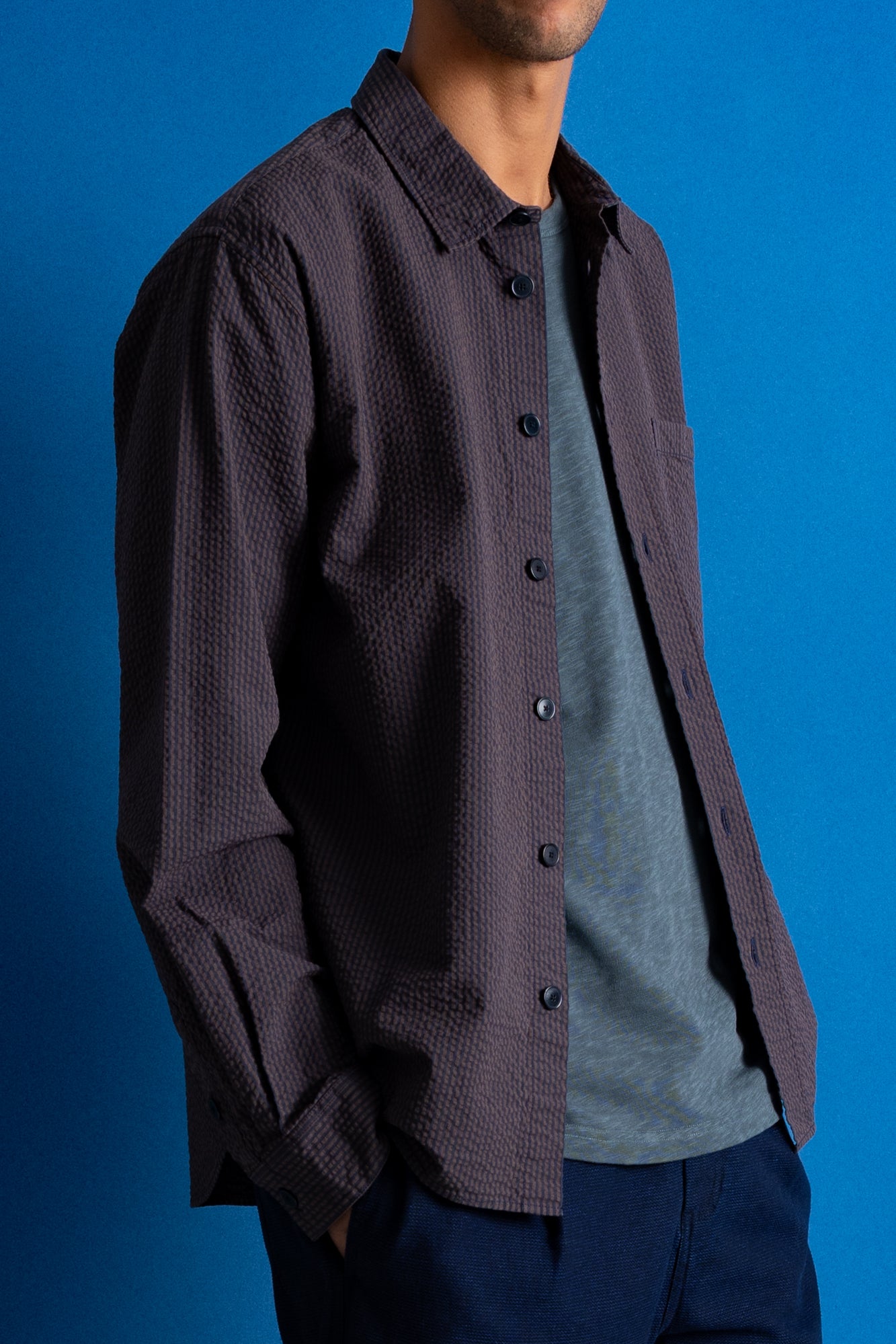 A man wearing a dark textured button-up shirt over a heather gray t-shirt and navy pants, posed against a solid blue studio background.