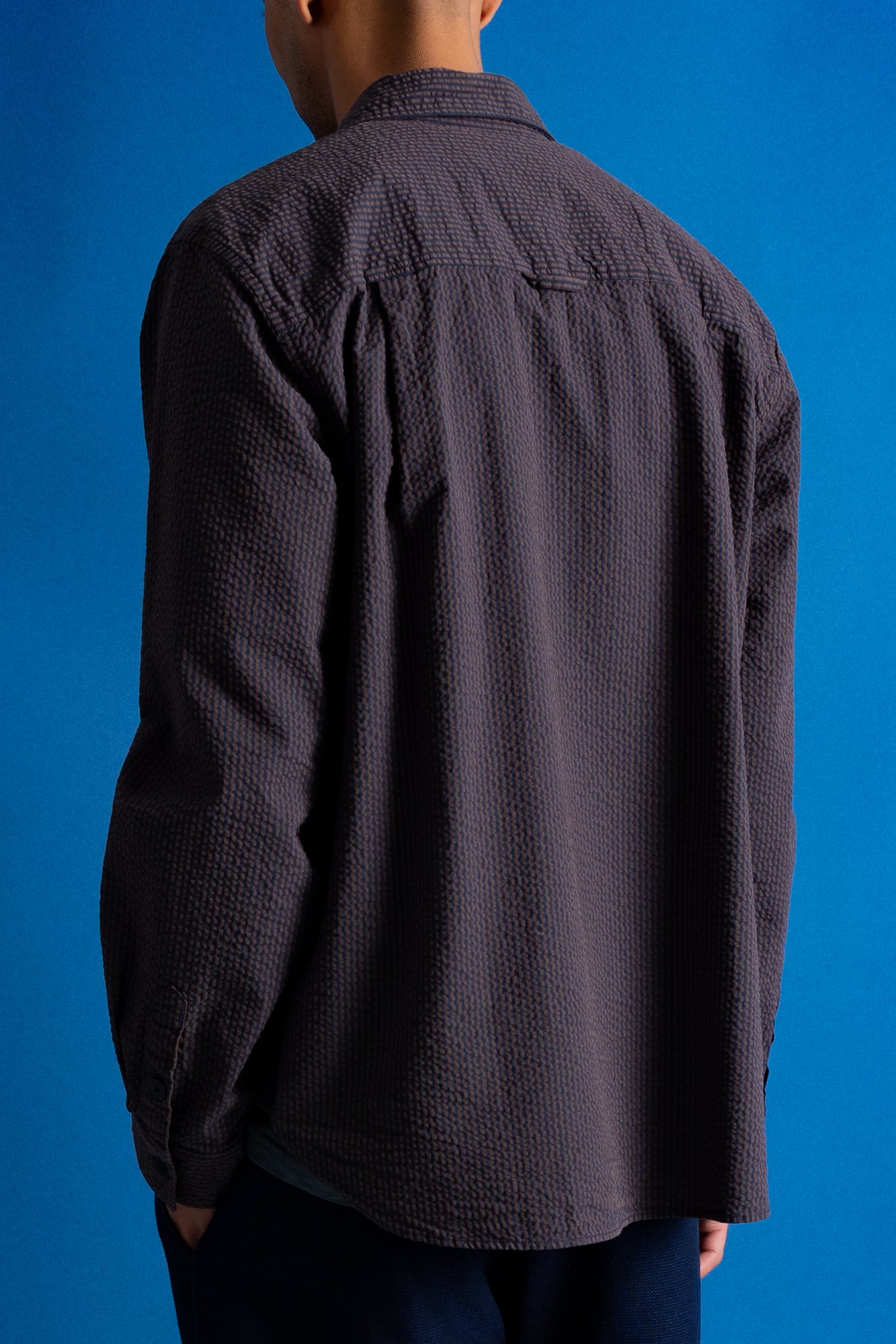 A man viewed from the back wearing a dark charcoal textured button-down shirt against a solid blue background.