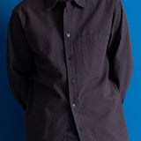 A man wearing a dark charcoal textured long-sleeve button-up shirt with a chest pocket, shown from the neck down against a solid blue background.