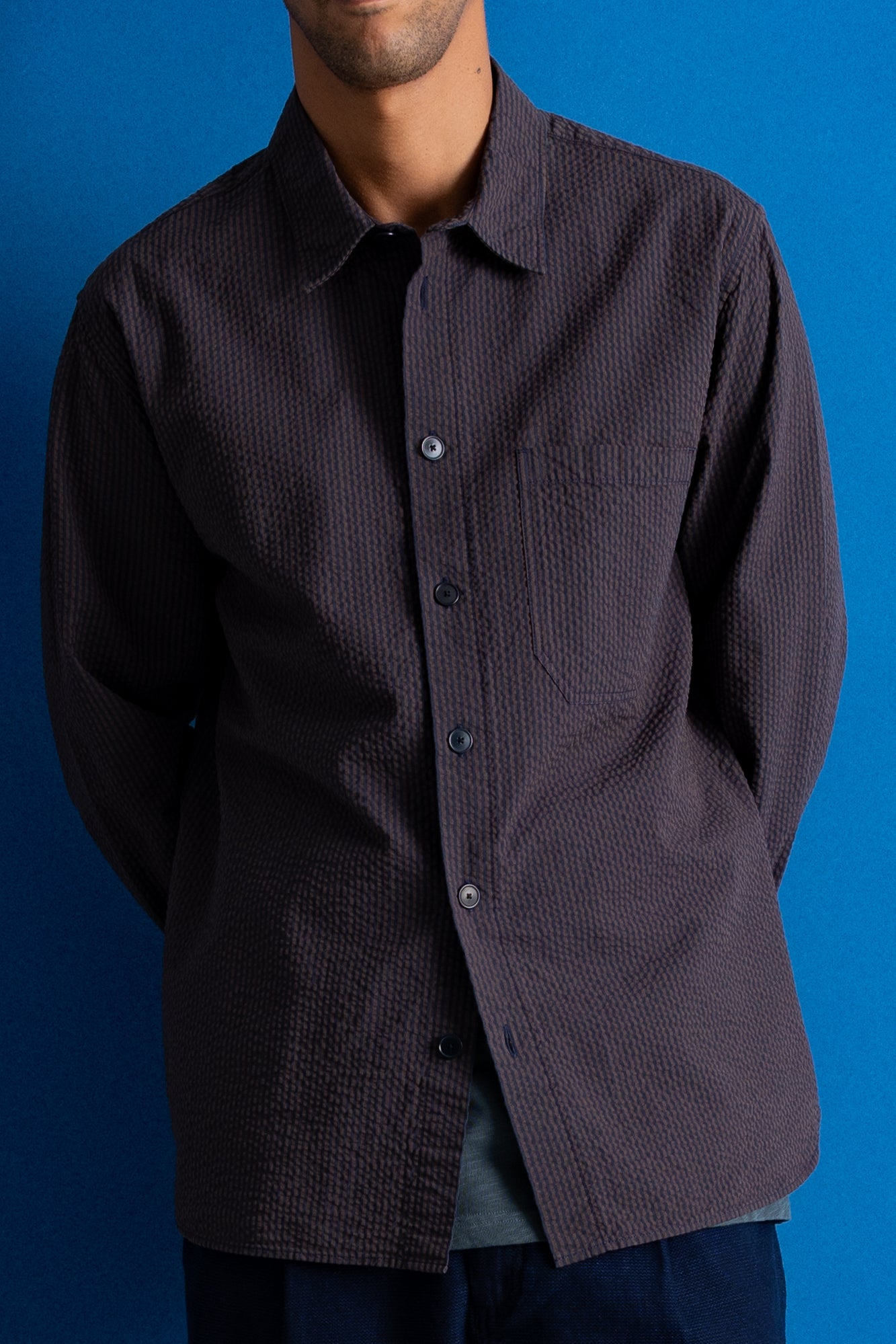 A man wearing a dark charcoal textured long-sleeve button-up shirt with a chest pocket, shown from the neck down against a solid blue background.