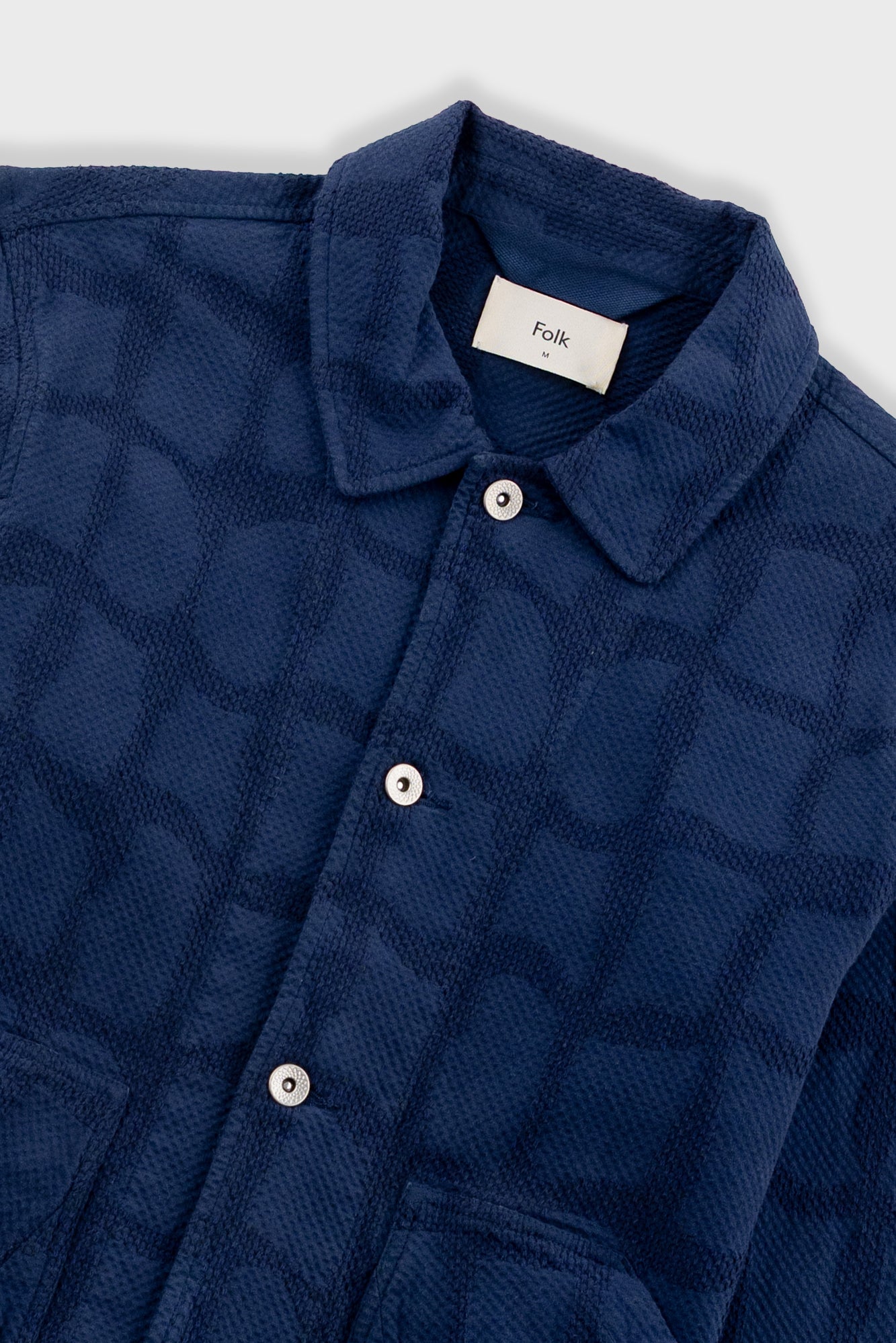 Close-up of a blue textured shirt with buttons and a visible brand label on a white background