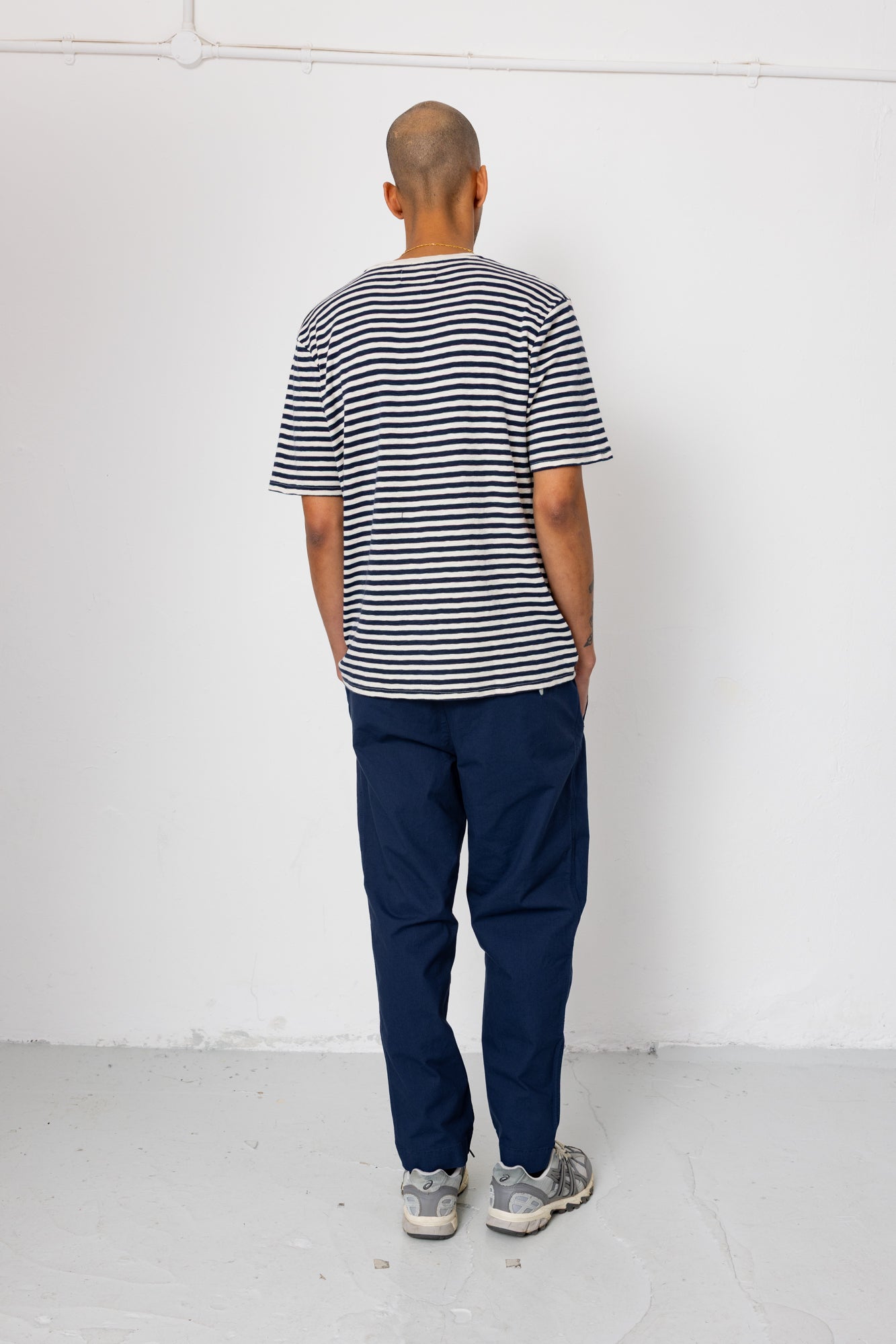 Man wearing a striped shirt and blue pants standing against a white wall.