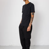 Man wearing a black t-shirt and pants standing against a white wall.