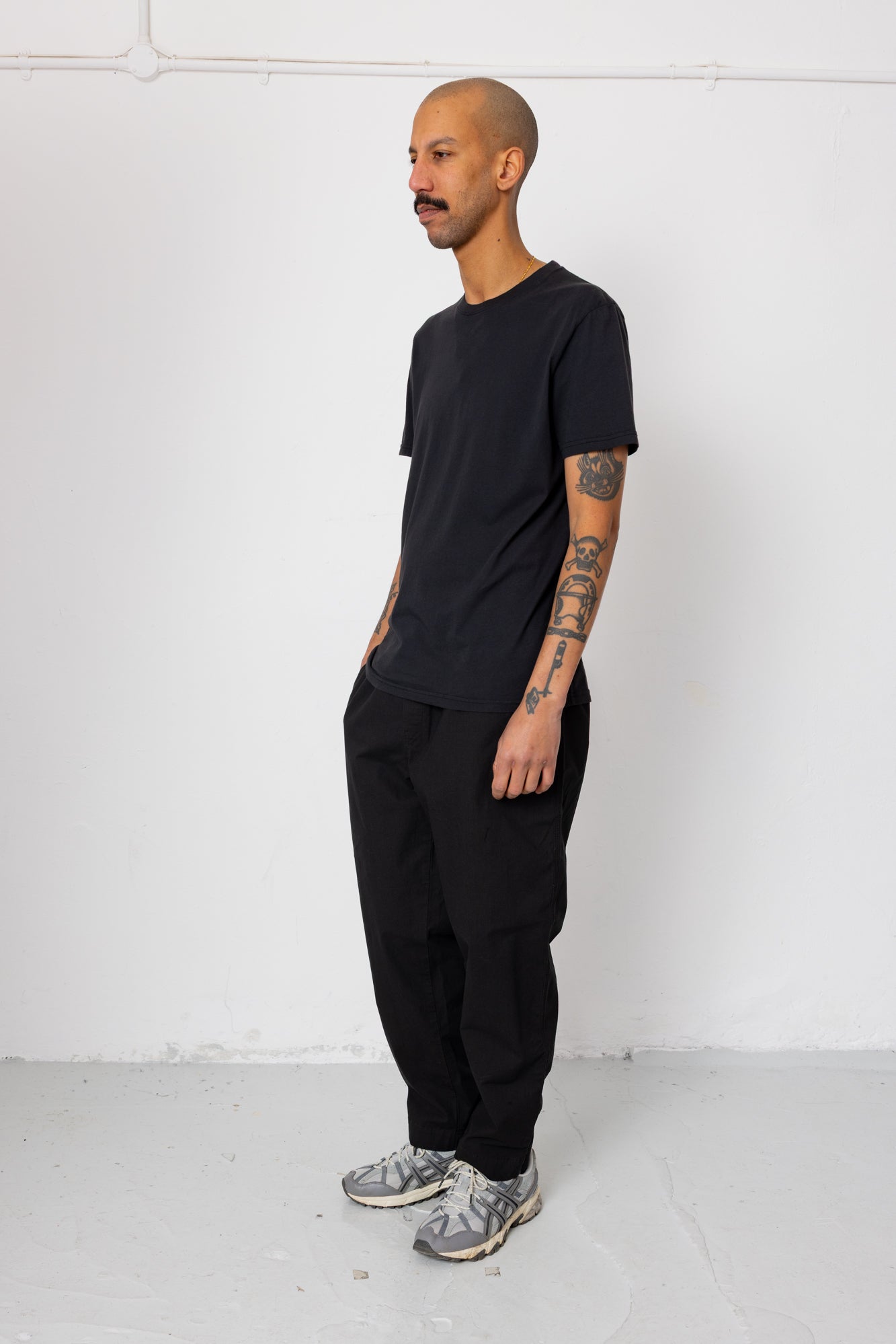 Man wearing a black t-shirt and pants standing against a white wall.