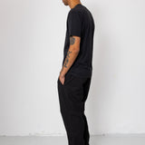 Man wearing a black t-shirt and pants standing against a white wall.