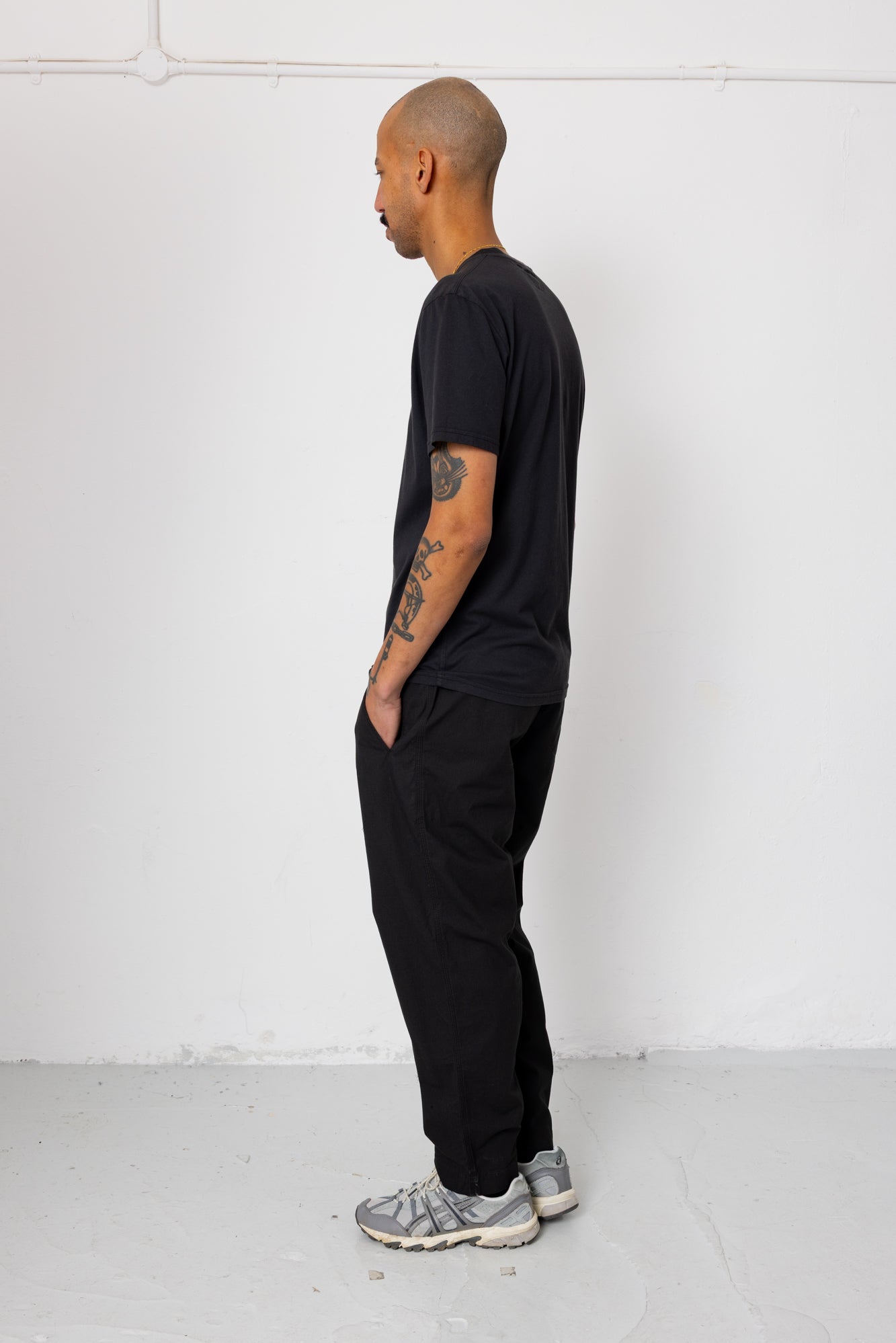 Man wearing a black t-shirt and pants standing against a white wall.