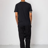 Person wearing a black t-shirt and pants standing against a white wall.