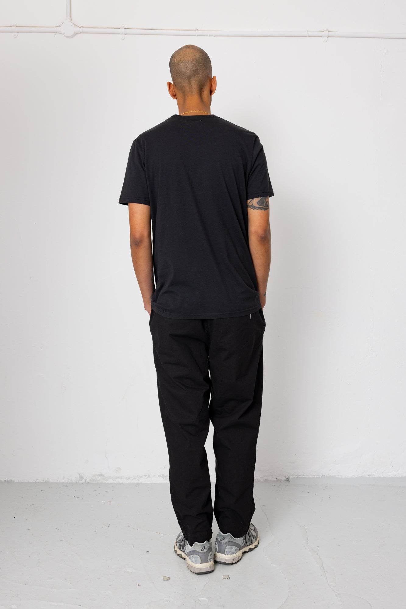 Person wearing a black t-shirt and pants standing against a white wall.