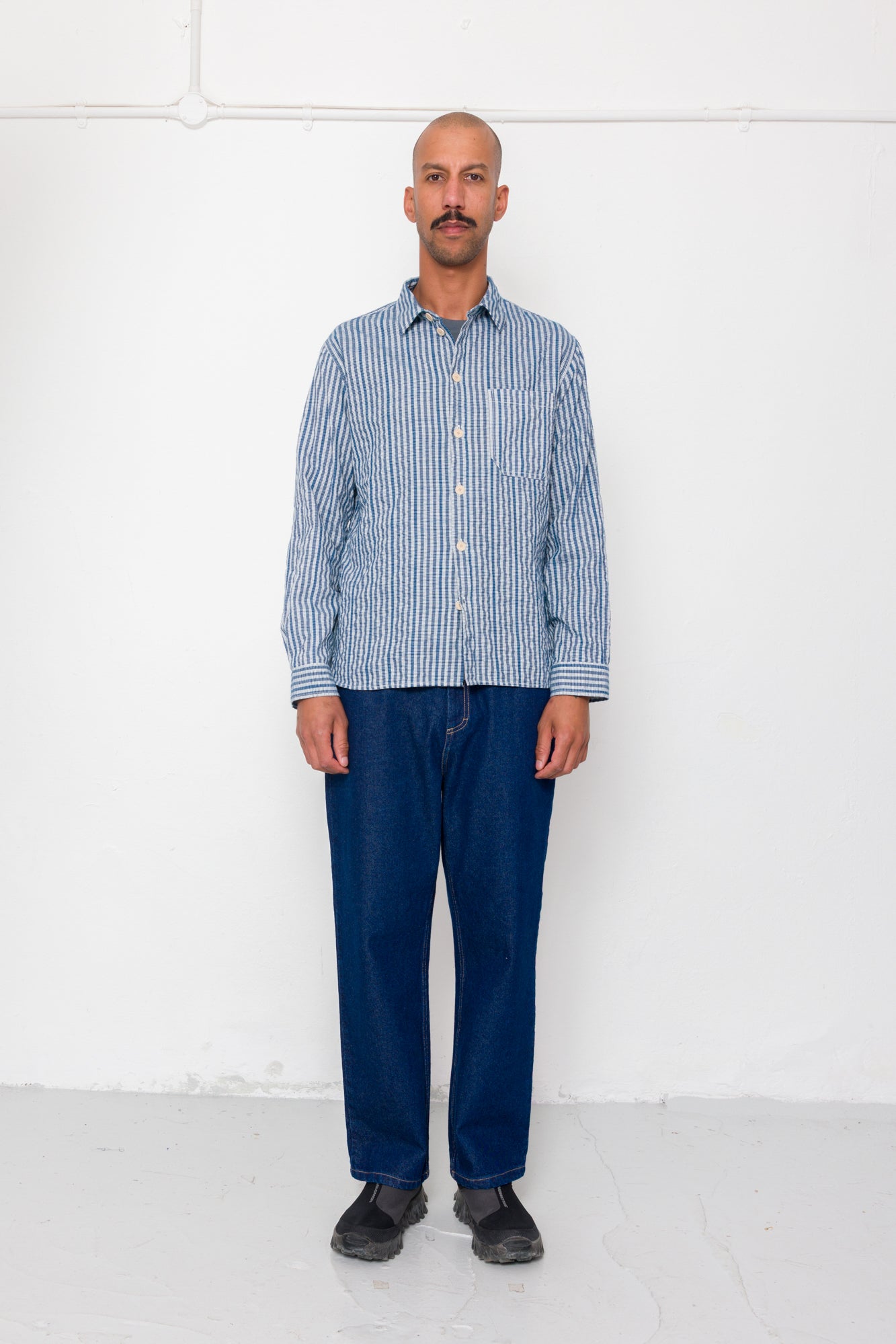 Man wearing a blue checkered shirt and dark pants against a white background