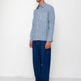 Man wearing a blue checkered shirt and dark jeans standing against a white wall.