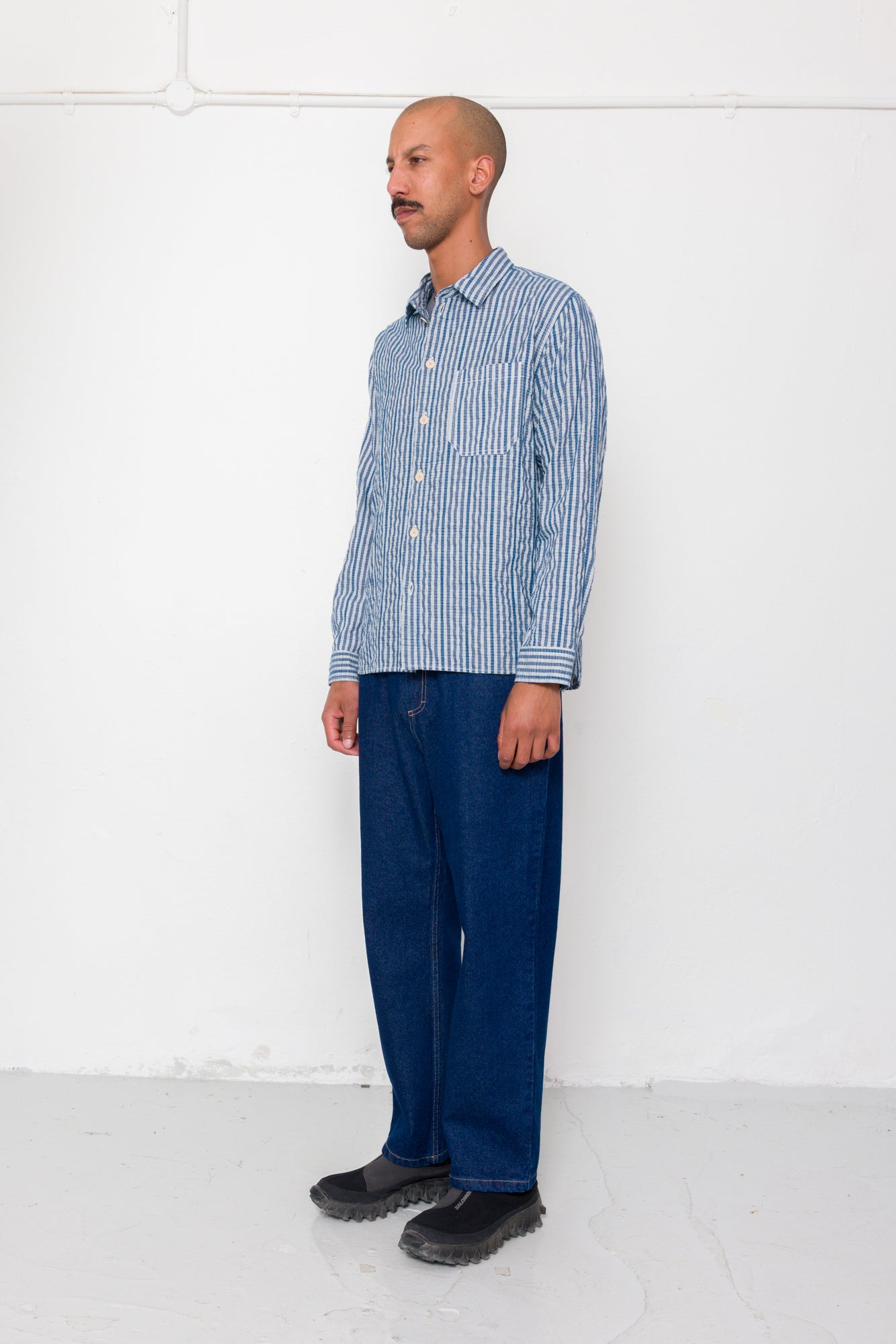 Man wearing a blue checkered shirt and dark jeans standing against a white wall.