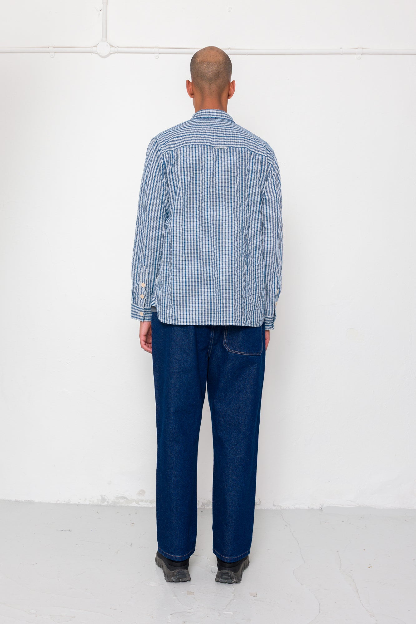 Person wearing a blue striped shirt and dark jeans standing against a white wall.