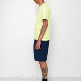 Man wearing a light yellow t-shirt and navy shorts standing against a white wall.