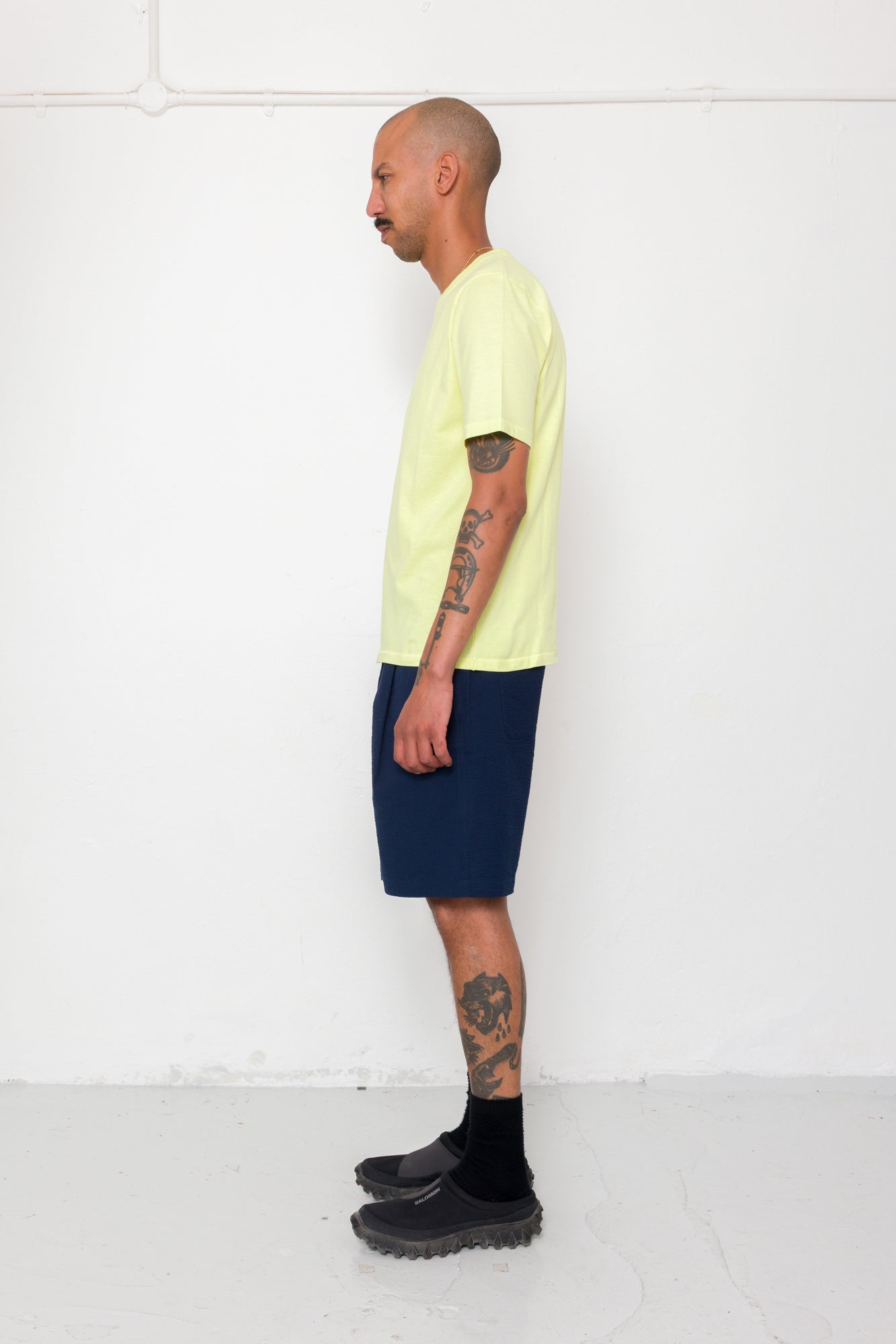 Man wearing a light yellow t-shirt and navy shorts standing against a white wall.
