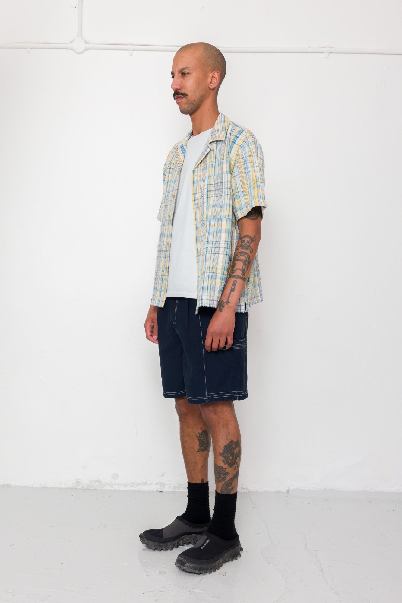 Man wearing a plaid shirt, white t-shirt, denim shorts, and black boots against a white background