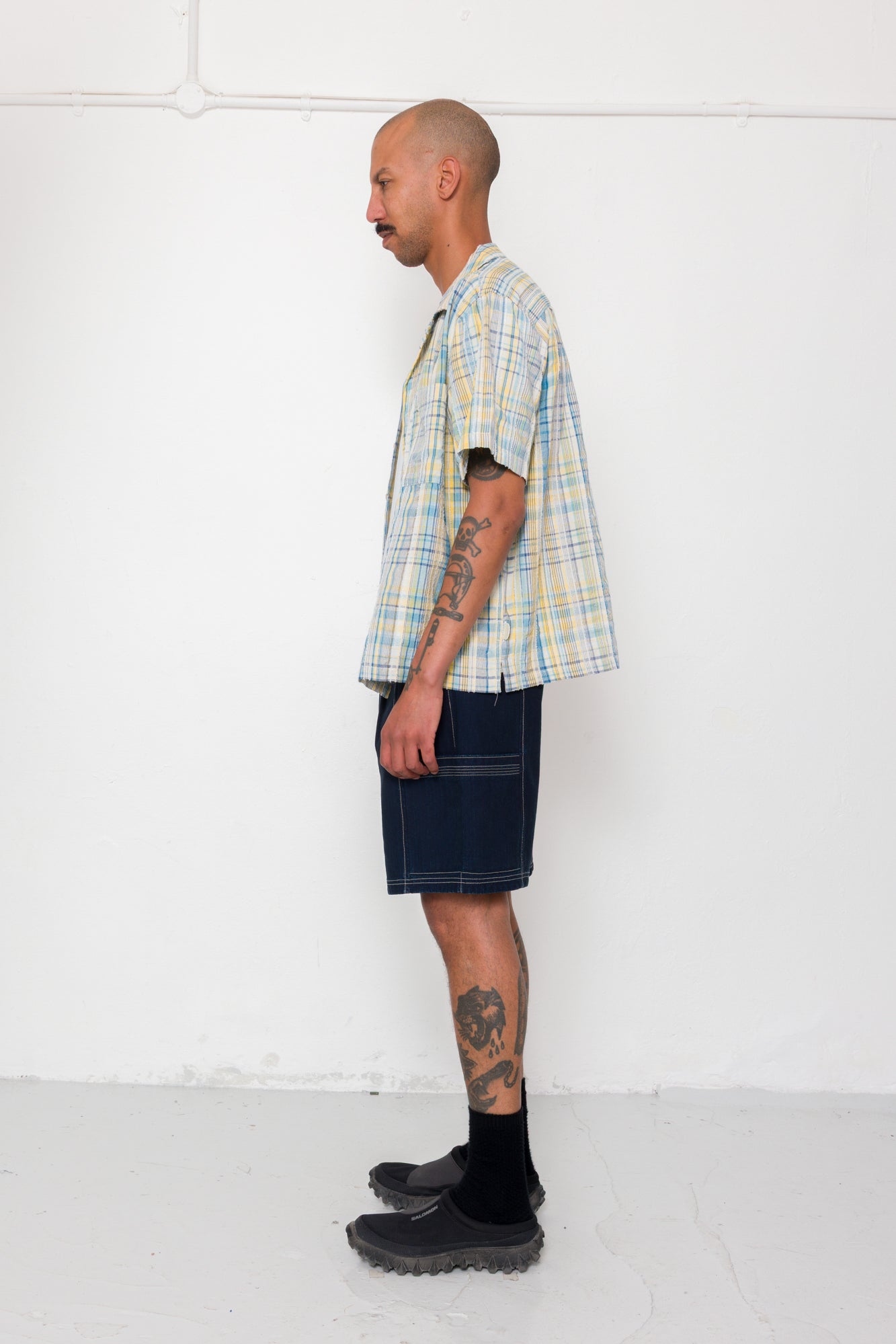 Man wearing a plaid shirt, dark shorts, and black shoes against a white background