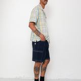 Man wearing a plaid shirt and dark shorts standing against a white wall.