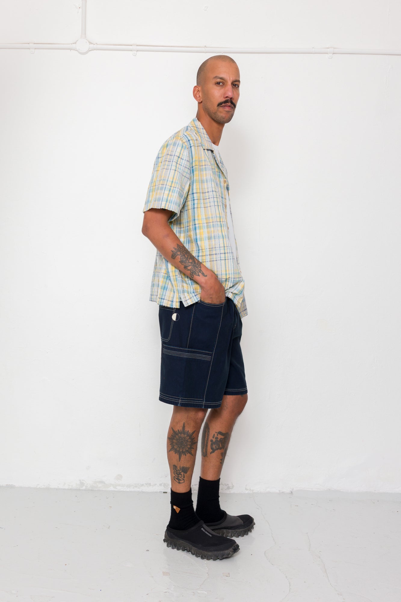 Man wearing a plaid shirt and dark shorts standing against a white wall.
