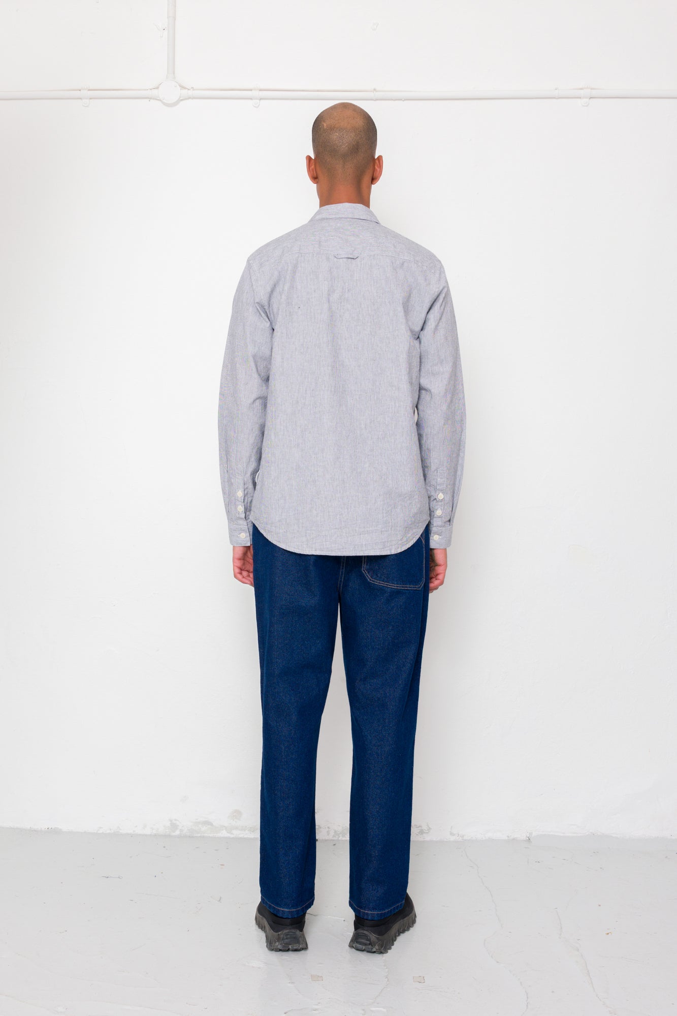 Man wearing a light gray shirt and dark blue jeans standing against a white wall.