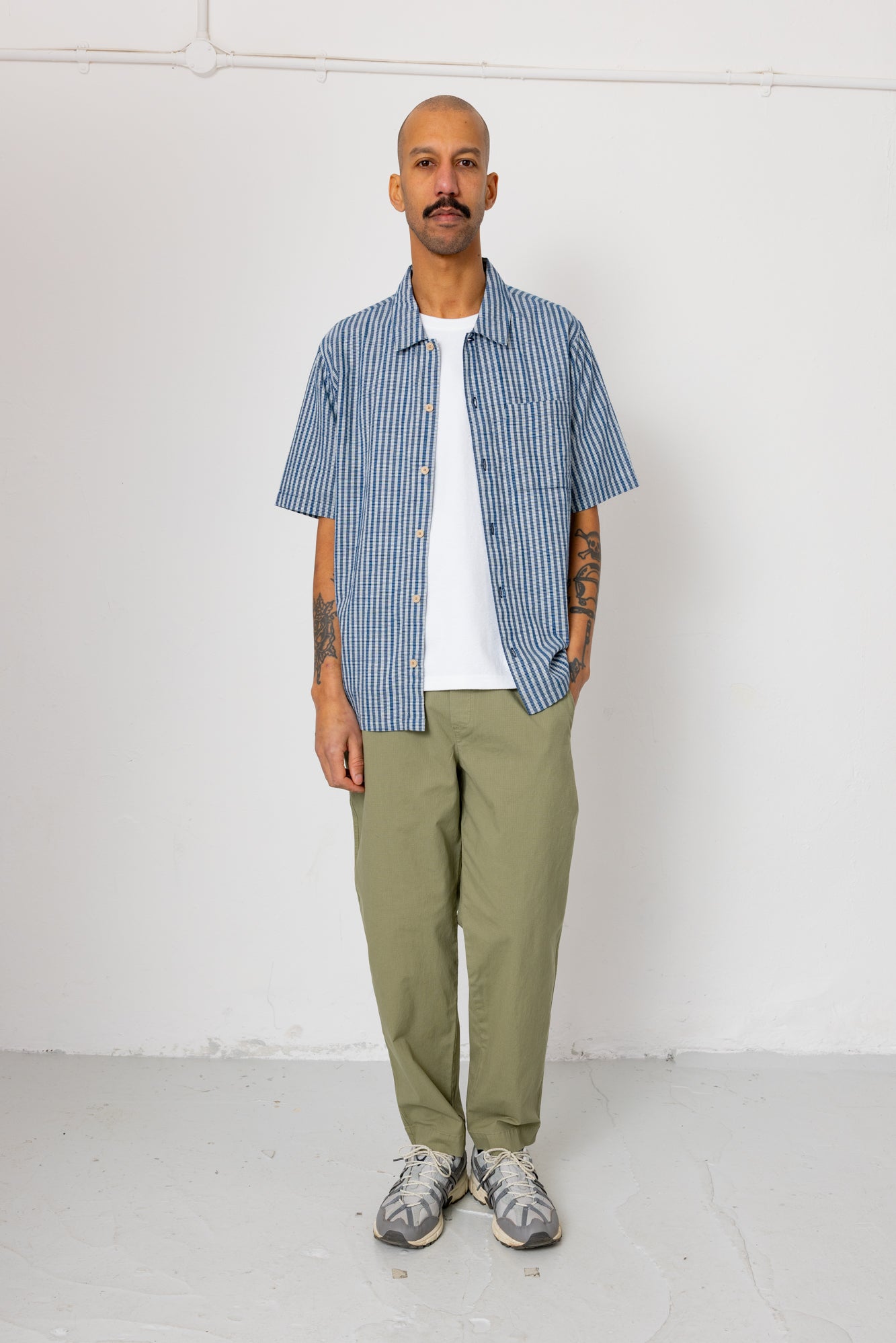 Man wearing a blue striped shirt, white t-shirt, green pants, and gray sneakers against a white background