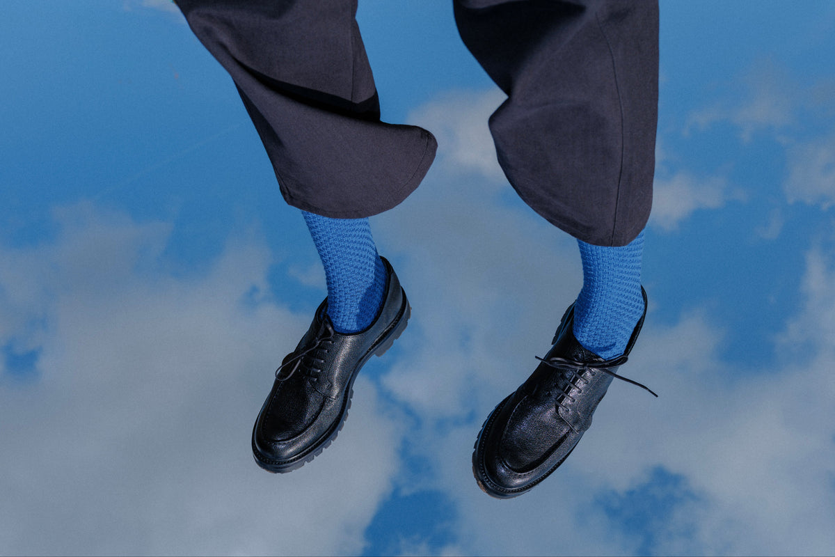 A person's legs dangling in the air, wearing loose black trousers, bright blue patterned socks, and black leather lace-up shoes against a reflective blue sky with clouds.