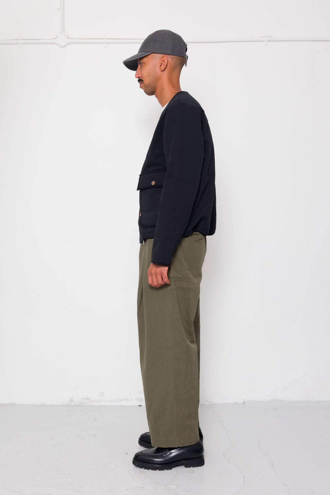 Man wearing a black quilted jacket, green pants, and a cap against a white background