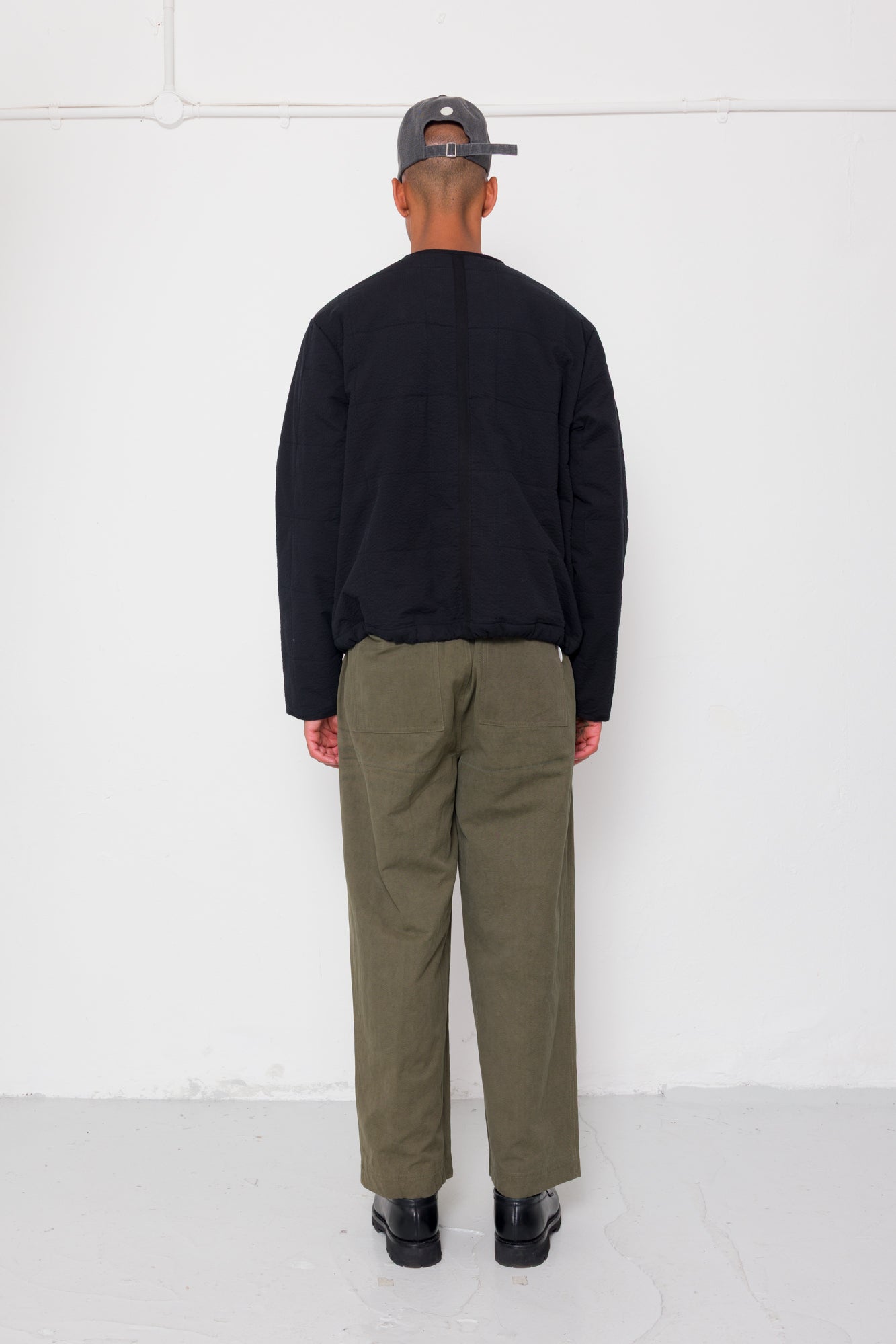 Man wearing a black quilted jacket, green pants, and a cap against a white background
