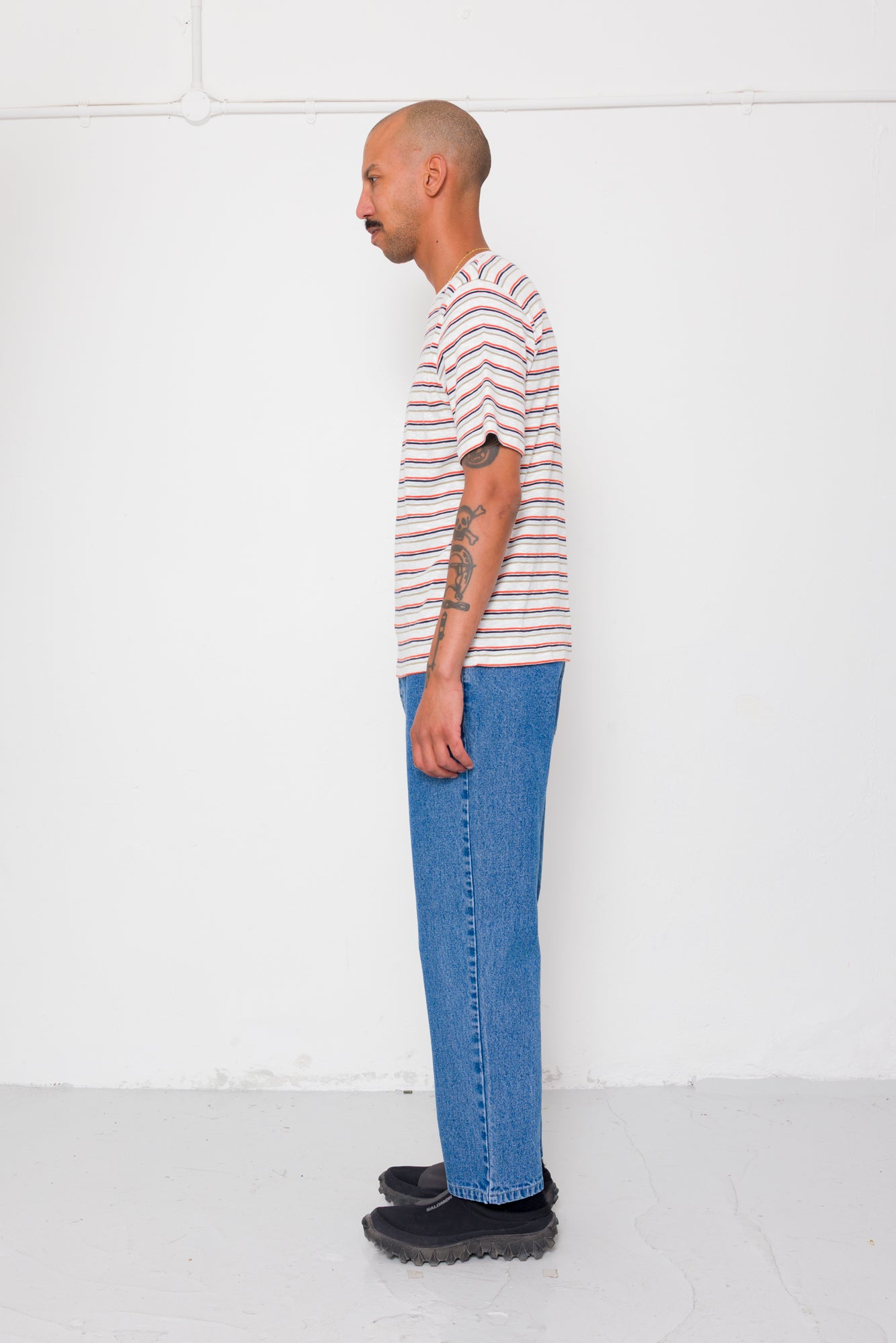 Man wearing a striped shirt and blue jeans standing against a white wall.