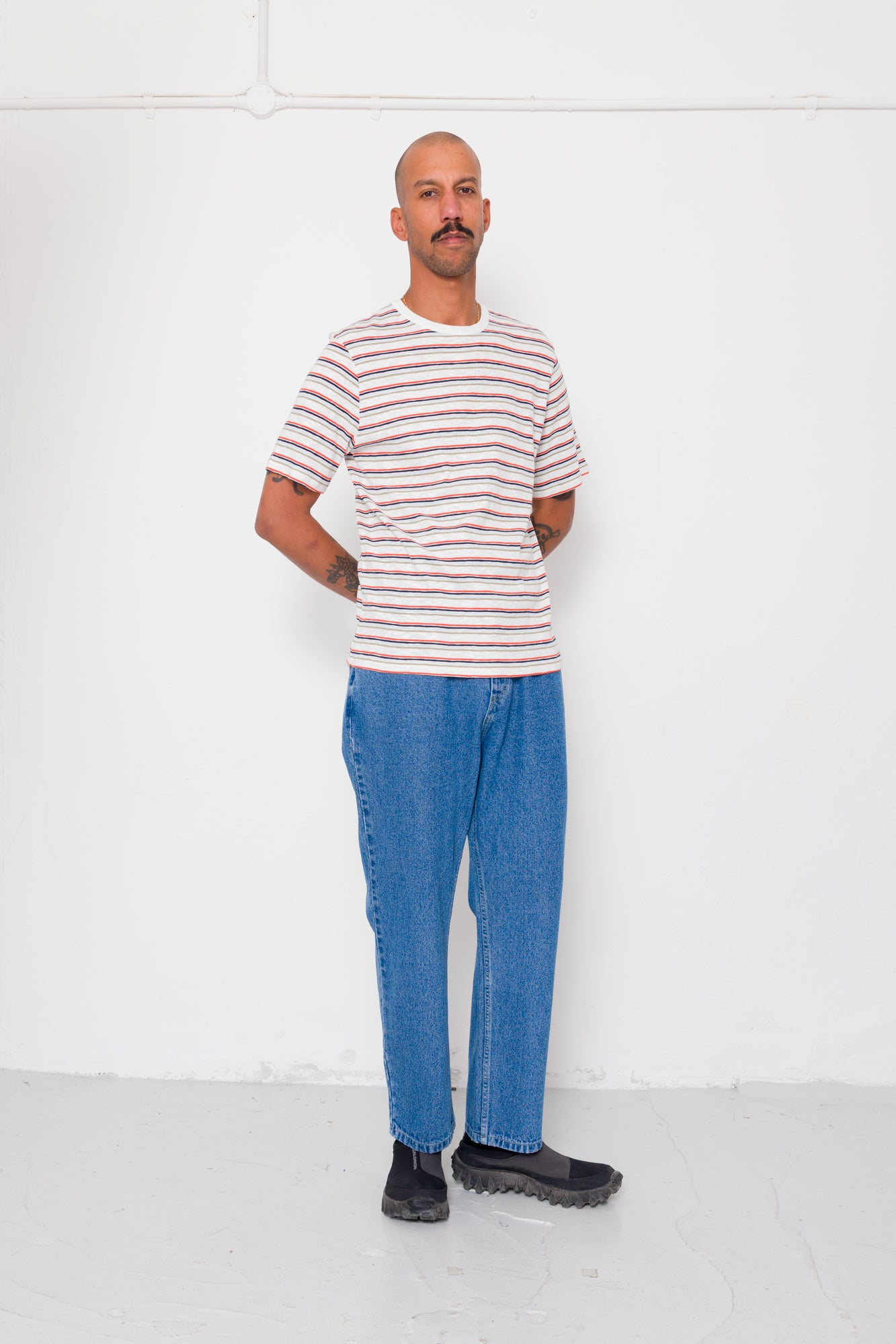 Man wearing a striped shirt and blue jeans standing against a white wall.