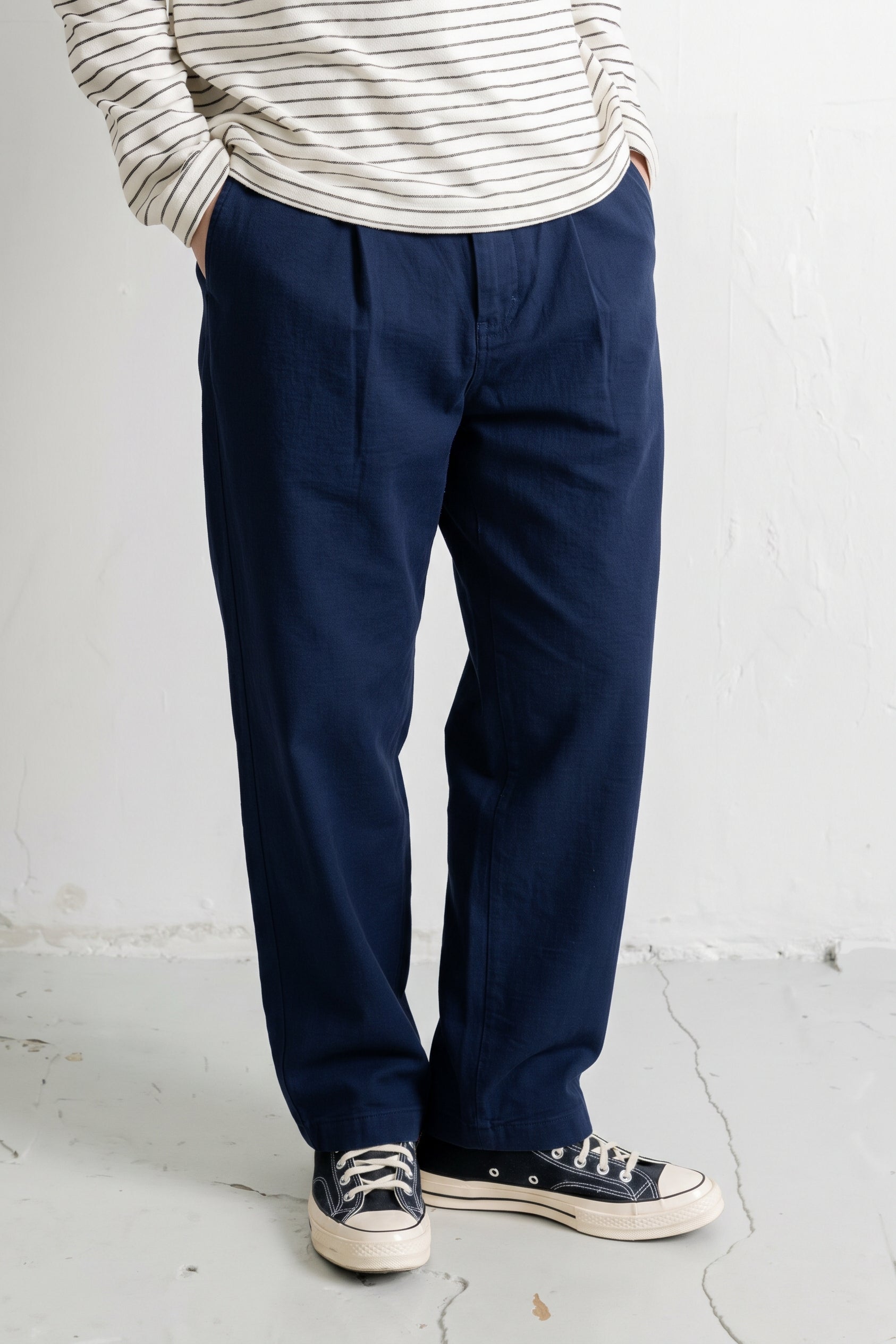 A person wearing navy blue relaxed-fit trousers, a cream and navy striped long-sleeve shirt, and navy canvas sneakers, standing against a white textured wall.
