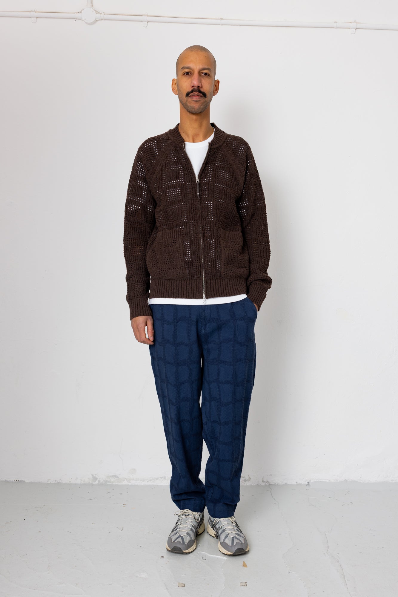 Man wearing a brown cardigan, white shirt, blue checkered pants, and gray shoes against a white background