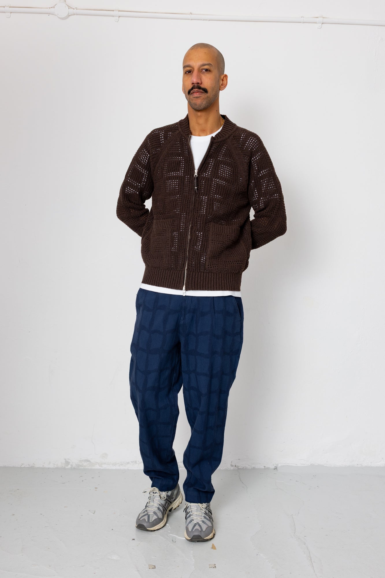 Man wearing a brown patterned sweater and blue pants against a white background