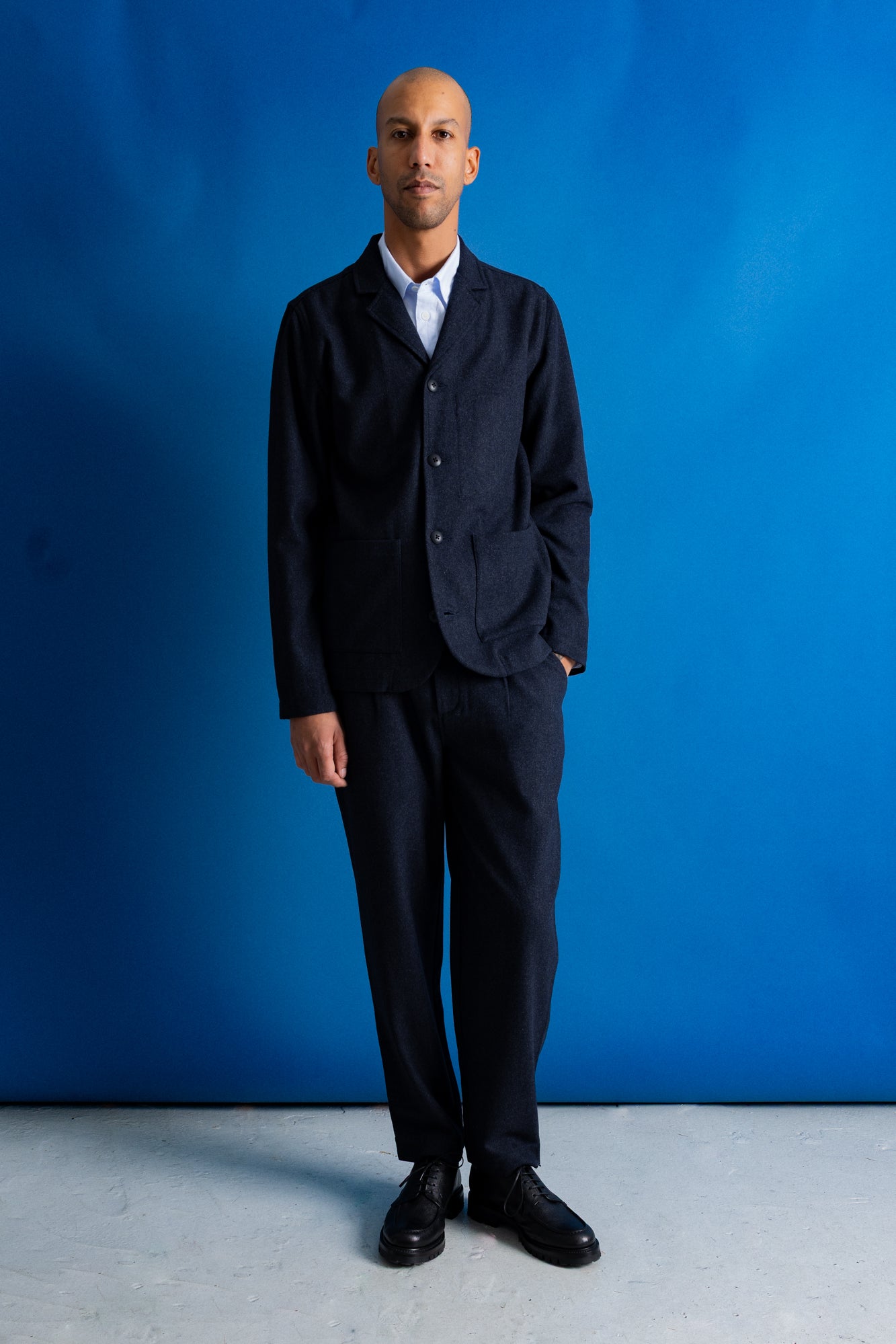 A man wearing a dark navy blue three-piece suit with a white collared shirt and black dress shoes, standing against a vibrant blue studio backdrop.