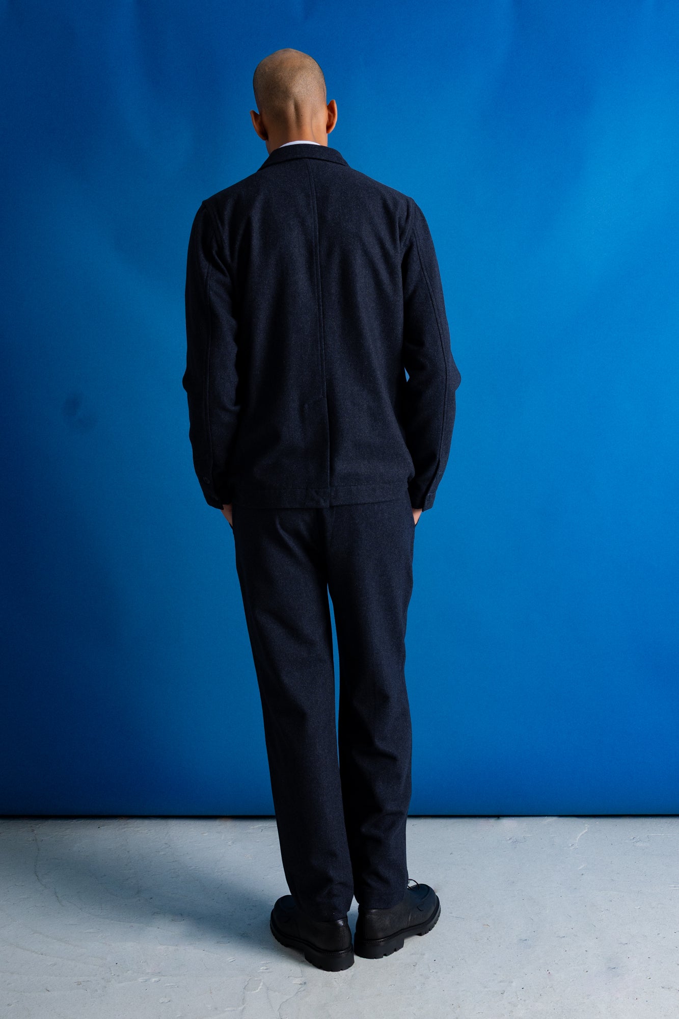 A man viewed from behind wearing a dark navy blue jacket and matching trousers, standing against a vibrant blue studio backdrop.