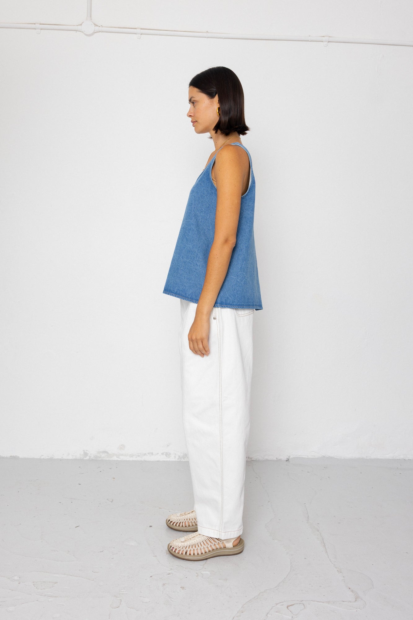 Woman wearing a denim tank top and white pants on a plain background