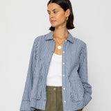 Woman wearing a blue striped shirt over a white top and green pants against a white background