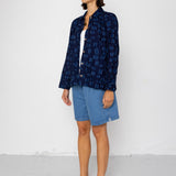 Woman wearing a dark blue patterned shirt over a white top with denim shorts, standing against a white background.