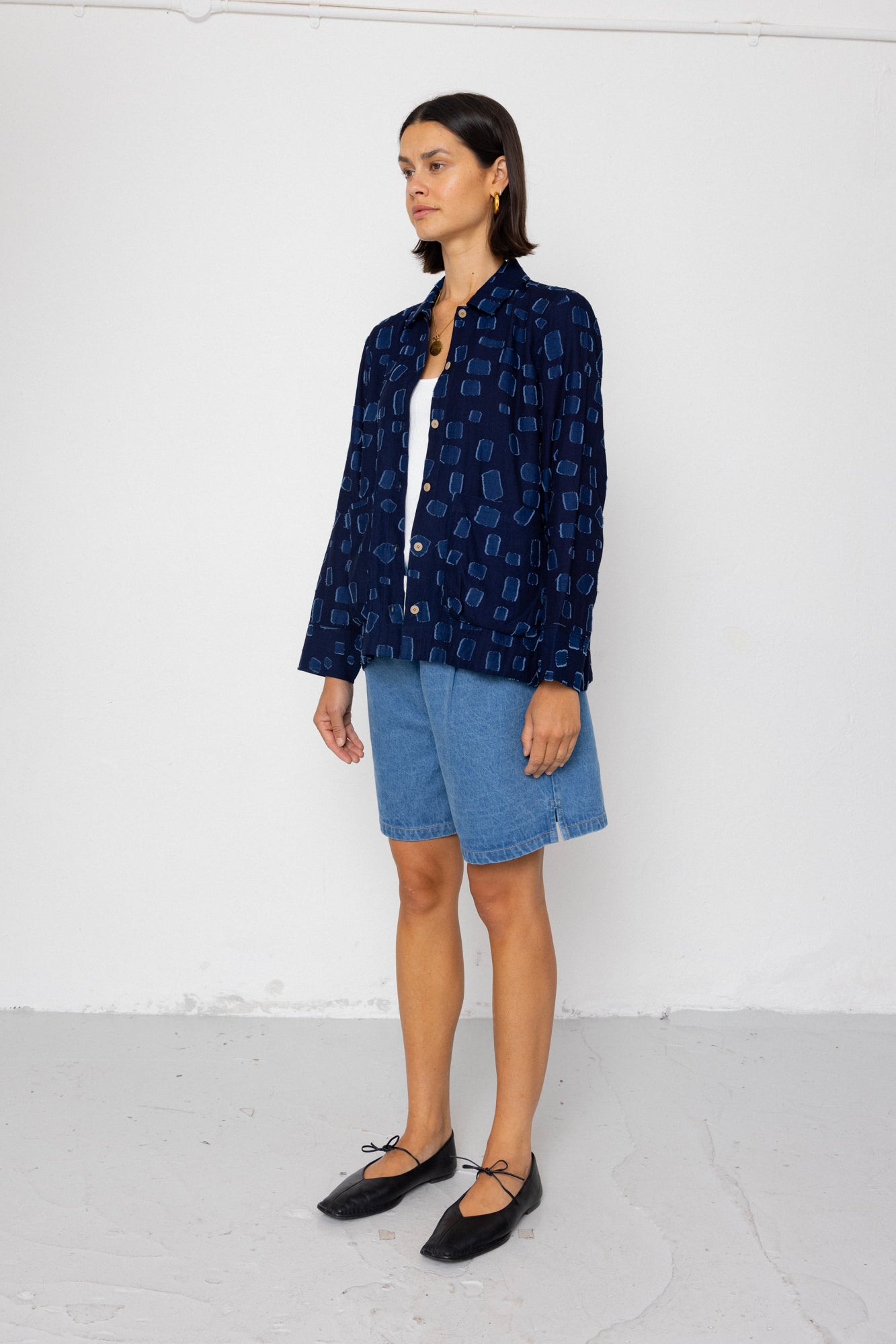 Woman wearing a dark blue patterned shirt over a white top with denim shorts, standing against a white background.