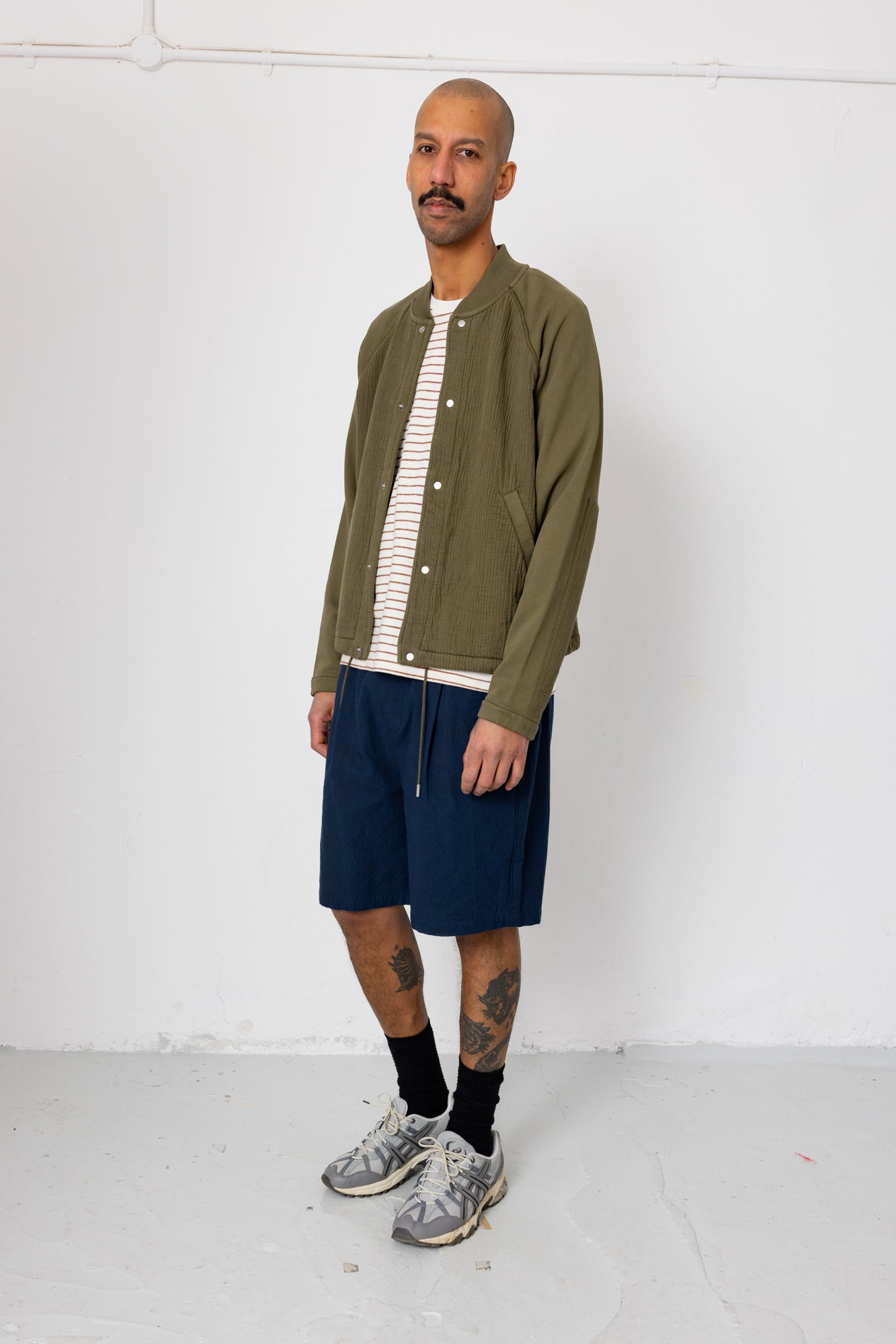 Man wearing a green jacket, striped shirt, navy shorts, and gray sneakers against a white background