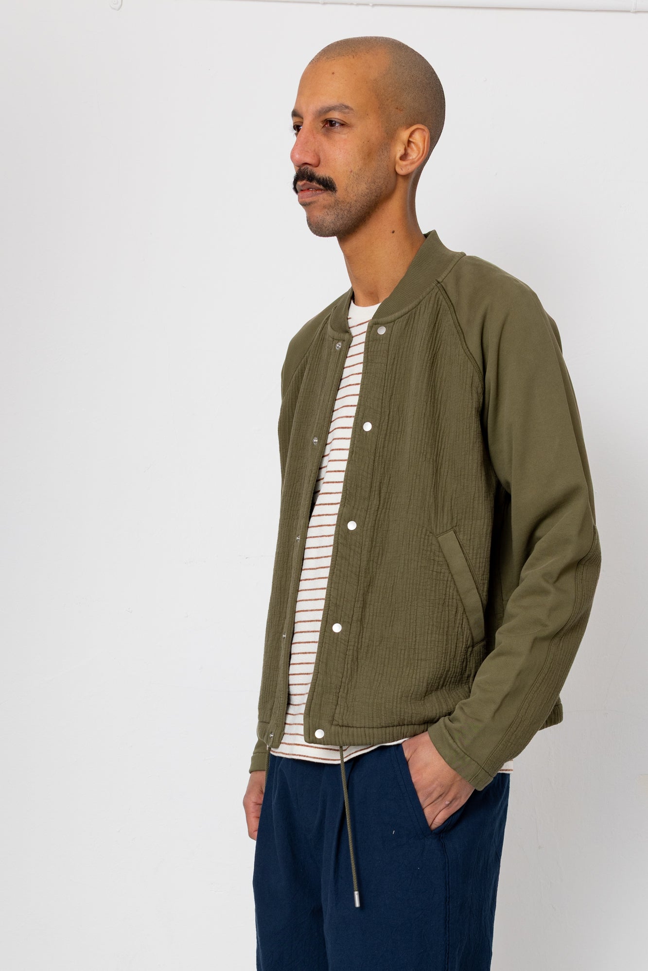 Man wearing a green jacket over a striped shirt with blue pants against a white background
