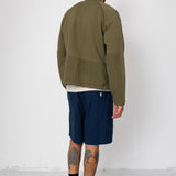 Man wearing an olive green jacket and navy shorts standing against a white wall.