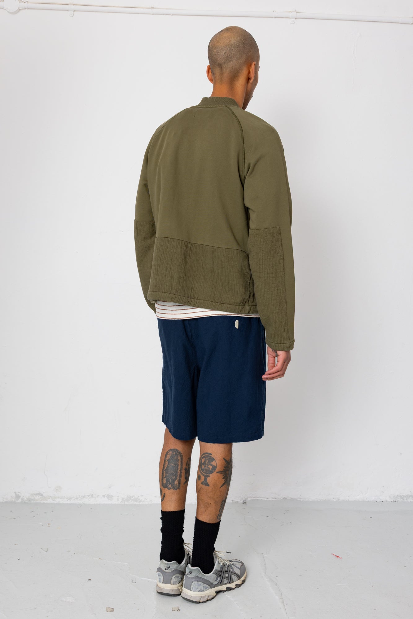 Man wearing an olive green jacket and navy shorts standing against a white wall.