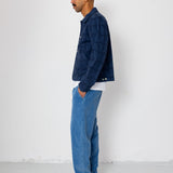 Man wearing a blue jacket and jeans standing against a white wall.